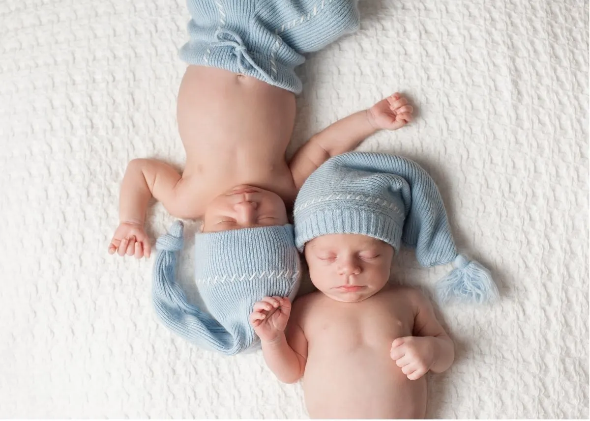 Twin boys with blue knit shorts and hats lay from opposite sides with their heads together.