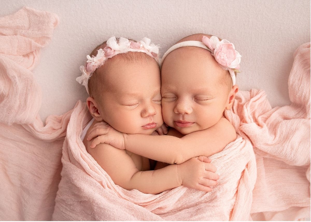 Twin baby girls with pink floral headbands embrace under a pink muslin blanket.